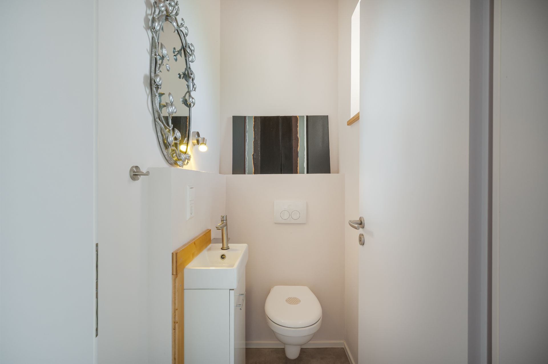 Kleines Badezimmer mit Wandtoilette, kleinem Waschbecken und rundem Spiegel.