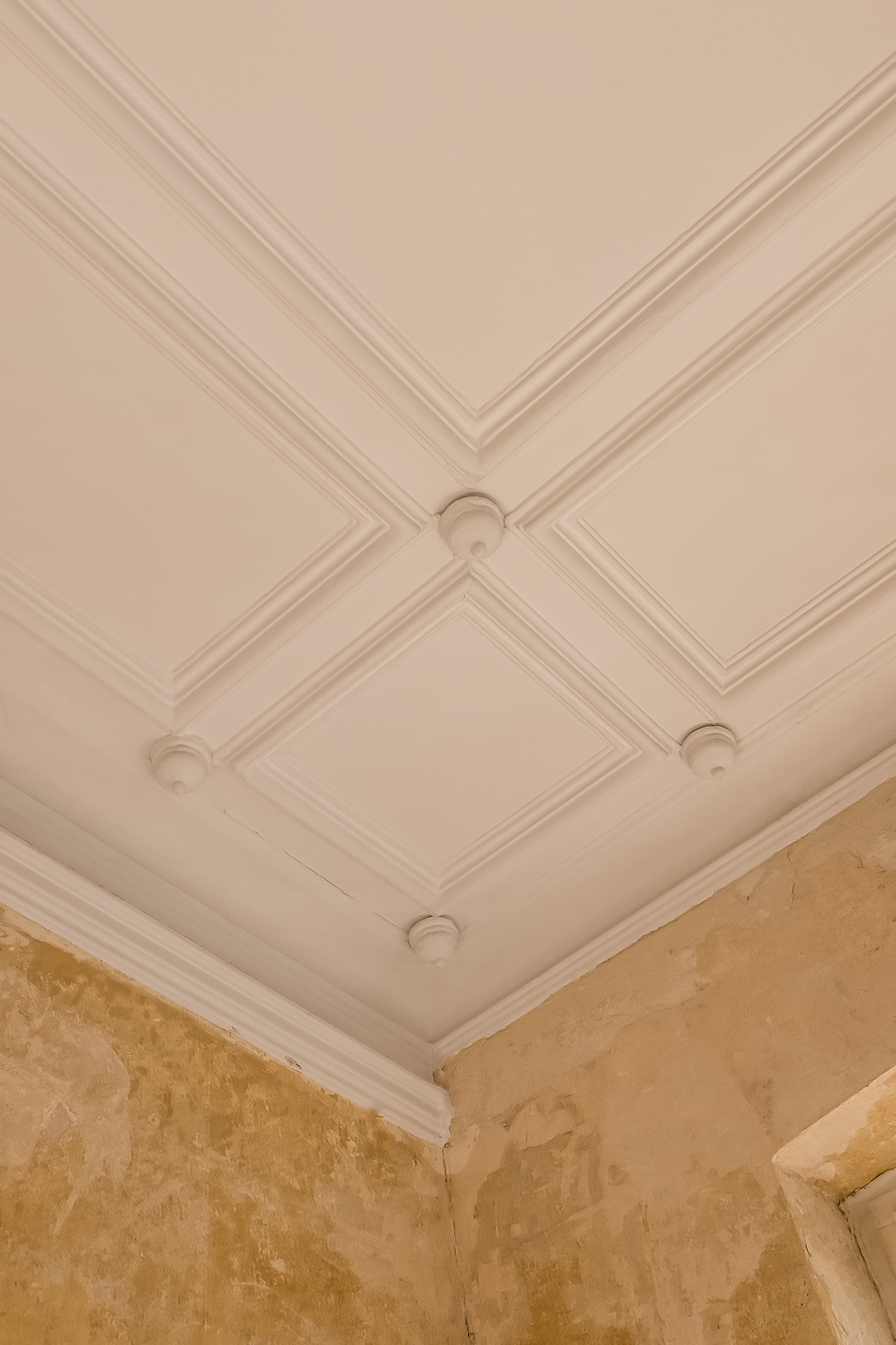 Weiße Stuckdecke mit geometrischem Muster und verzierten Ecken in einem Raum mit verputzter Wand.