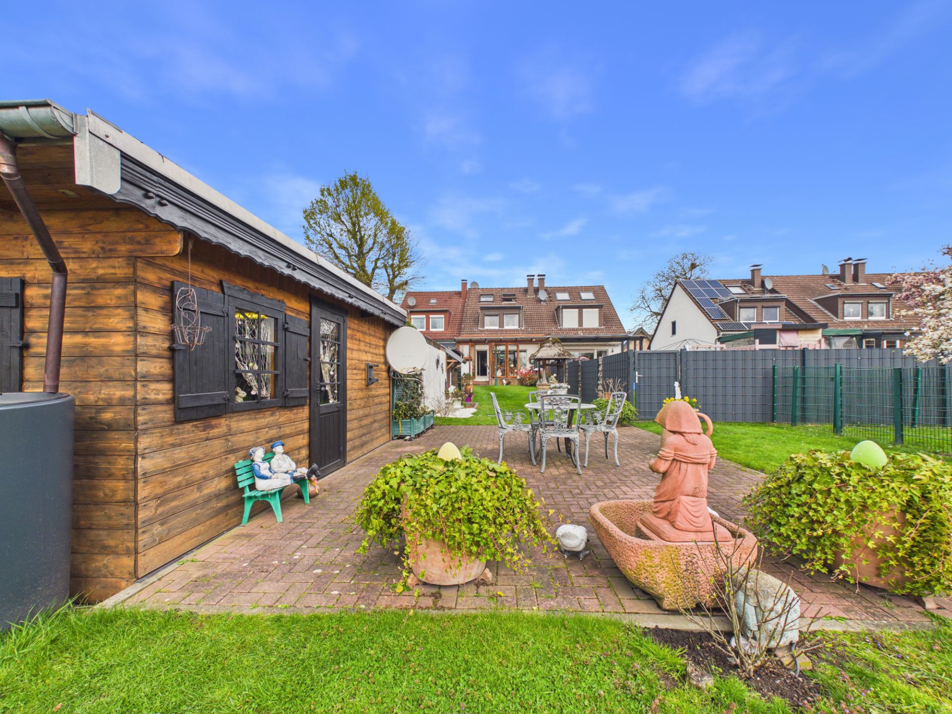 Garten mit Holzhaus, Sitzgruppe, Brunnenfigur und Pflanzenkübeln auf gepflasterter Terrasse.