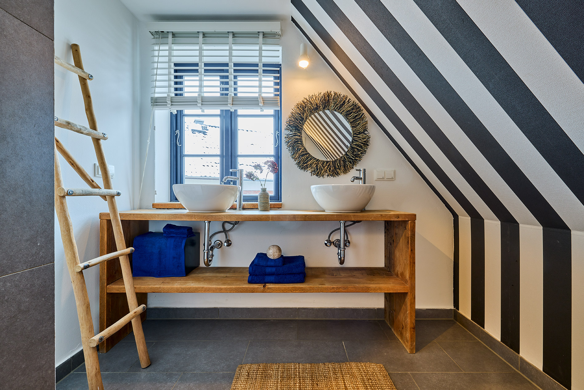 Modernes Bad mit zwei Waschbecken, Holzregal, gestreifter Wand und Leiterhandtuchhalter.