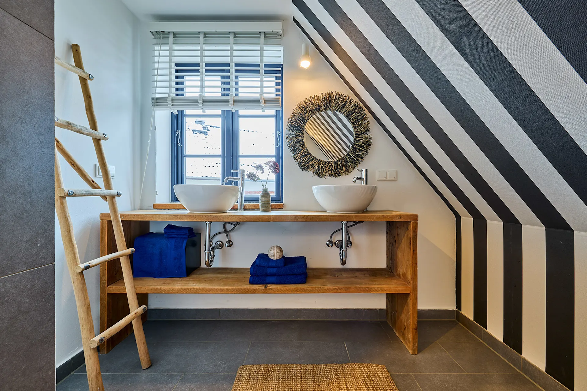 Modernes Bad mit zwei Waschbecken, Holzregal, gestreifter Wand und Leiterhandtuchhalter.