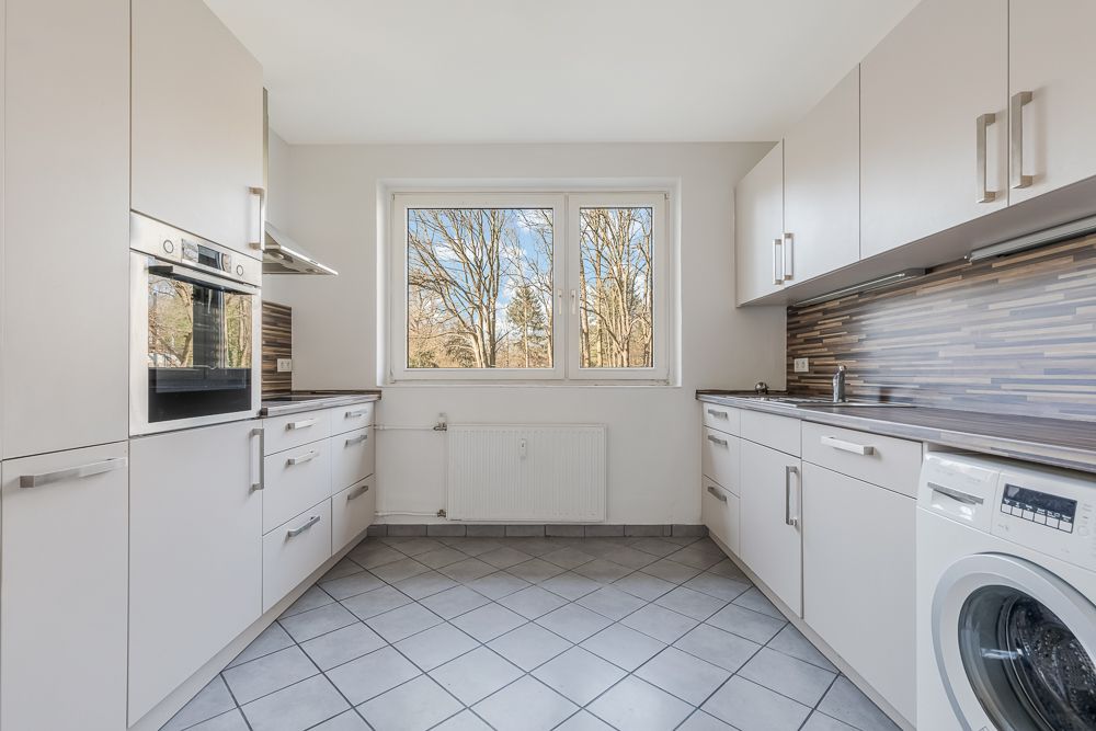 Helle Küche mit weißen Schränken, Backofen, Waschmaschine und Fenster mit Blick ins Freie.
