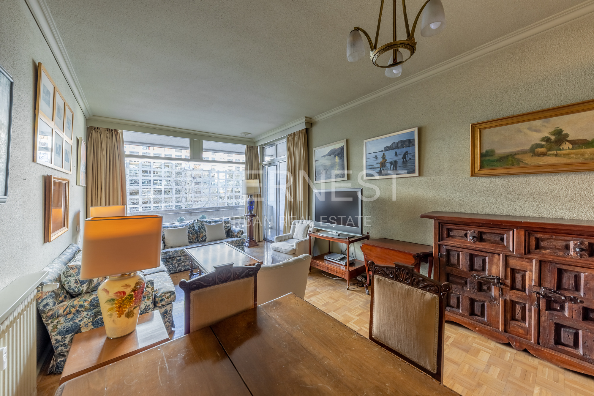 Helles Wohnzimmer mit Sofa, Sessel, TV, Holztisch und Kommode. Große Fenster mit Vorhängen.