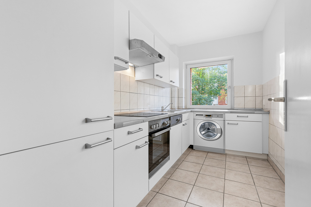 Moderne weiße Einbauküche mit Waschmaschine, Herd, Backofen und Fenster, Fliesenboden.