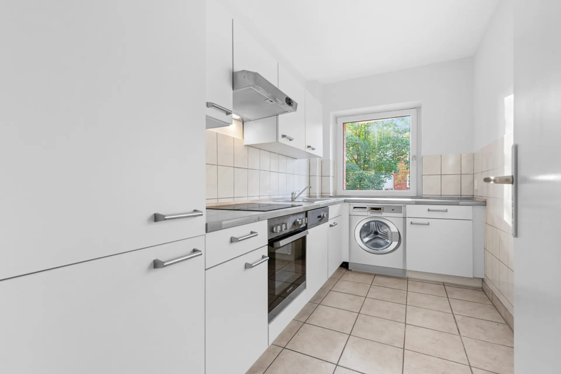 Moderne weiße Einbauküche mit Waschmaschine, Herd, Backofen und Fenster, Fliesenboden.
