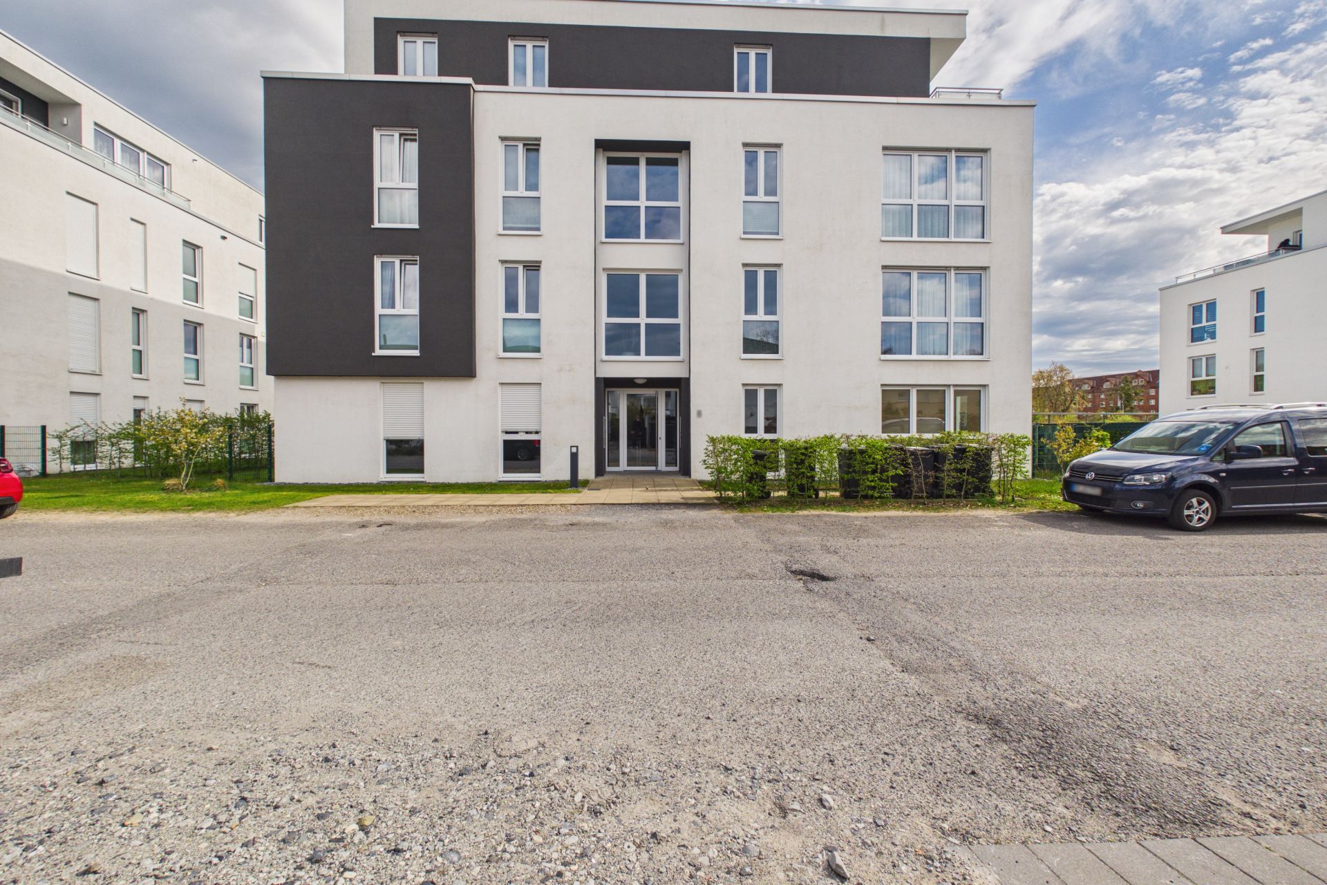 Modernes, mehrstöckiges Wohngebäude mit großen Fenstern und grauer Fassade, davor Straße und geparkte Autos.
