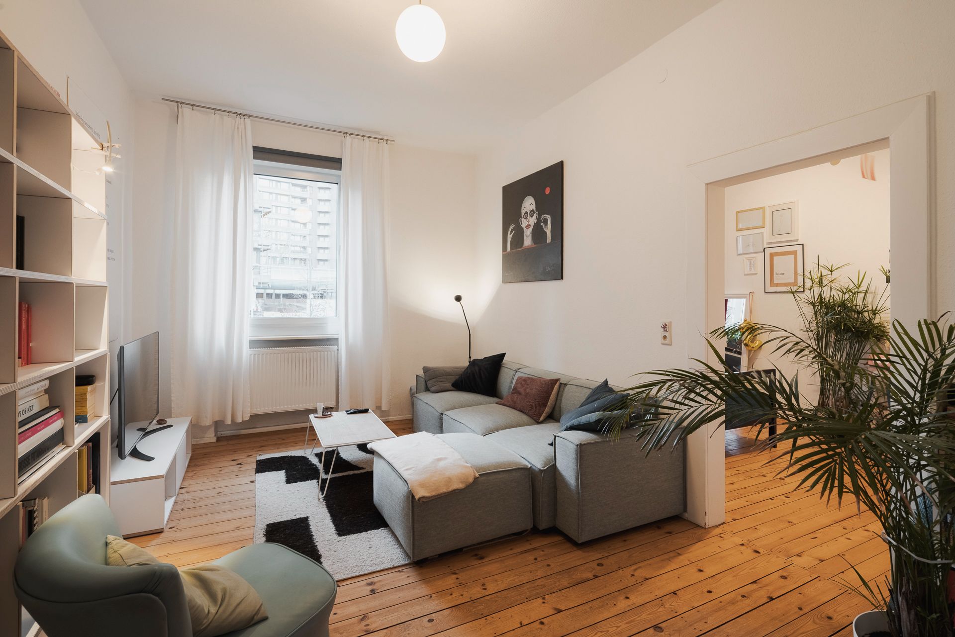 Wohnzimmer mit grauem Sofa, grünem Sessel, Bücherregal, Fernseher und großem Fenster mit weißen Vorhängen.