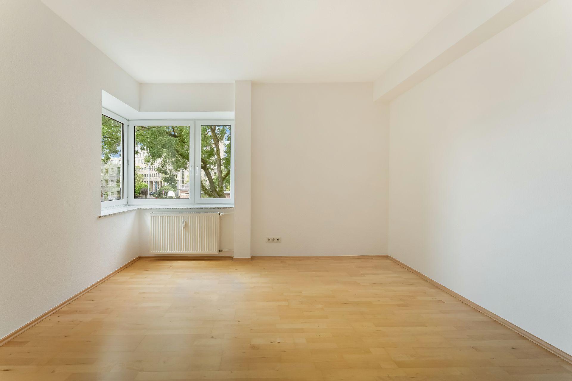 Leerer, heller Raum mit großem Eckfenster, Parkettboden und weiß gestrichenen Wänden.
