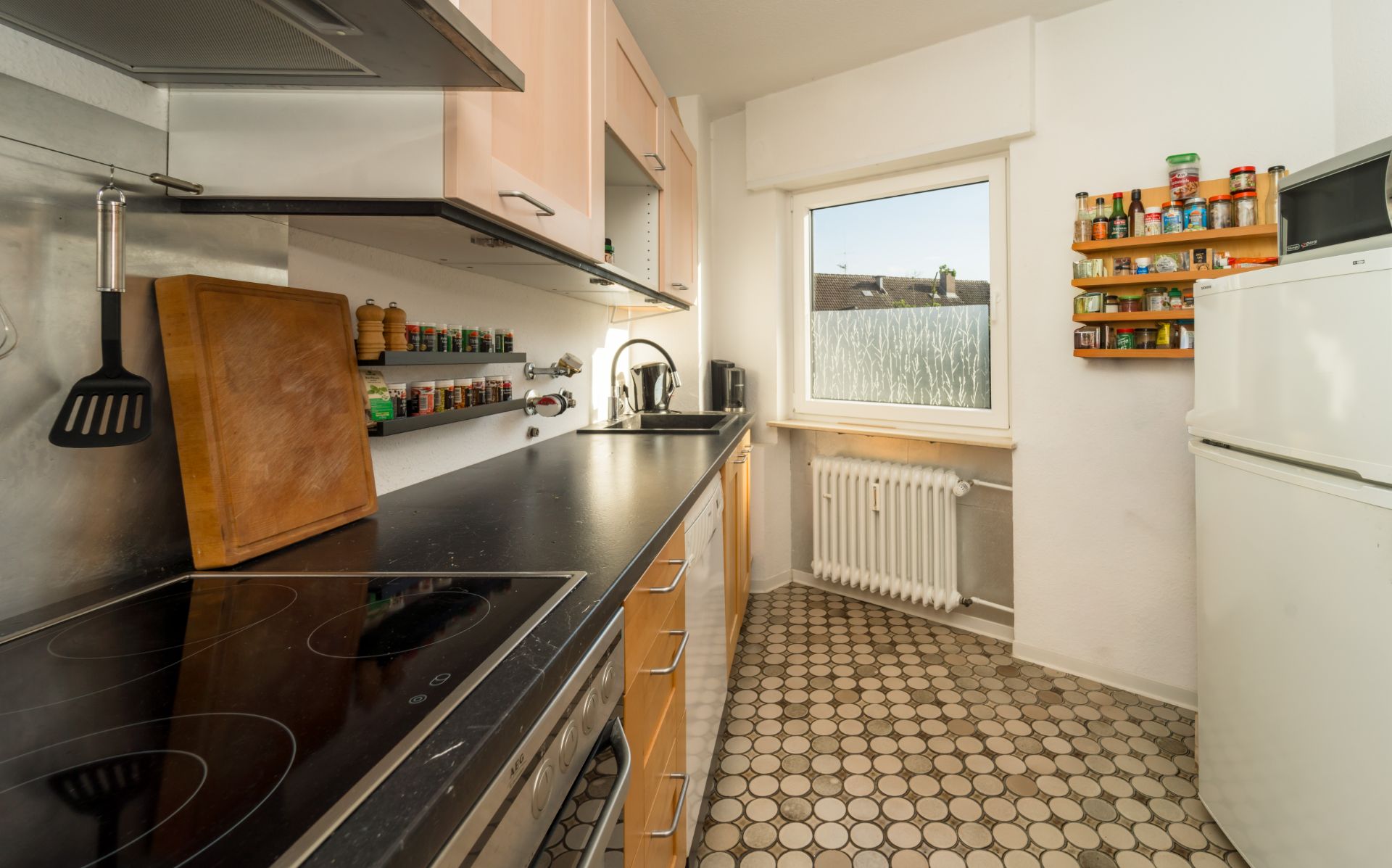 Schmale Küche mit Herd, Schneidebrett, Gewürzregalen, Fenster und Kühlschrank.