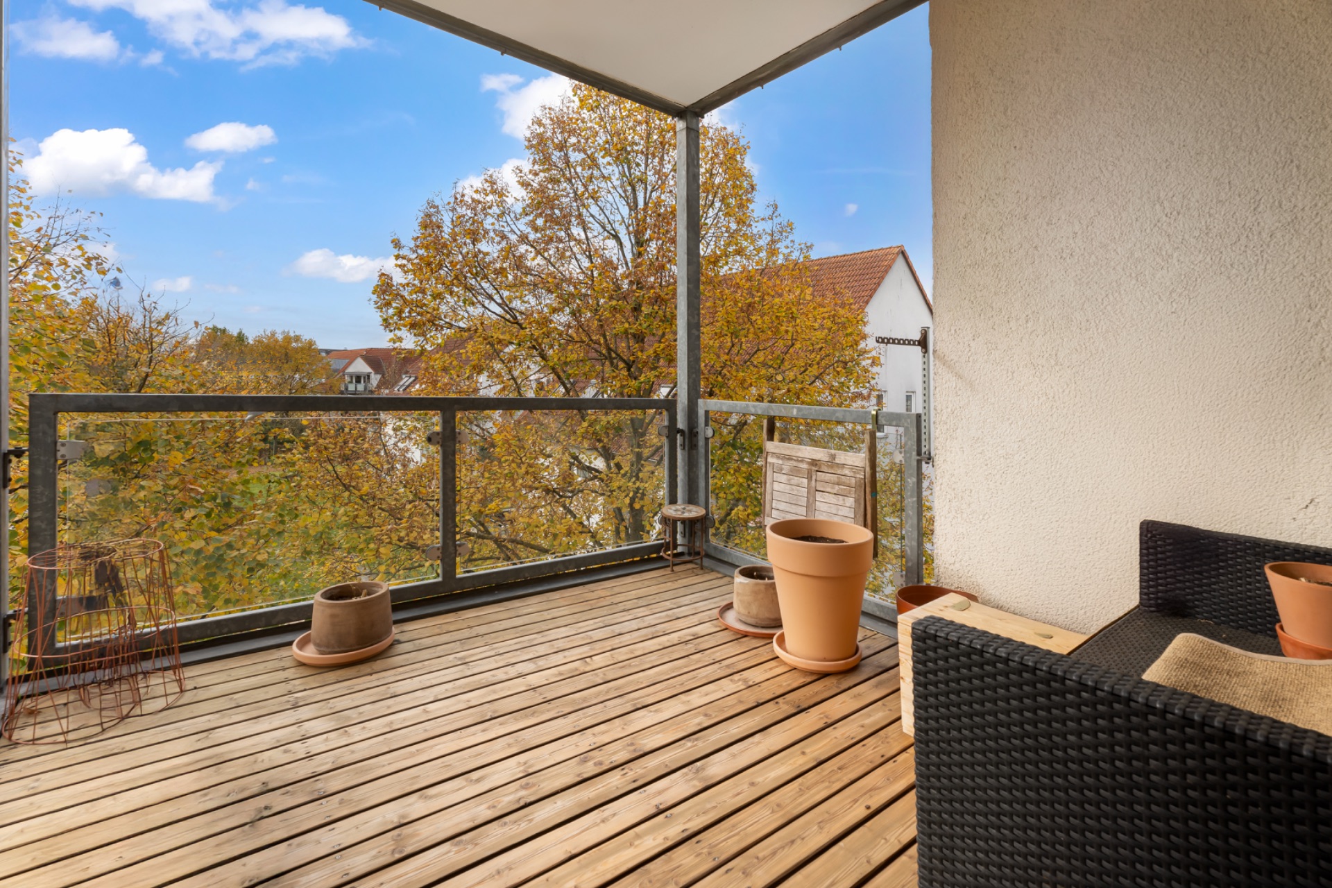 Holzbalkon mit Blumenkübeln, Stuhl und Sofa, Blick auf Bäume und angrenzende Häuser.
