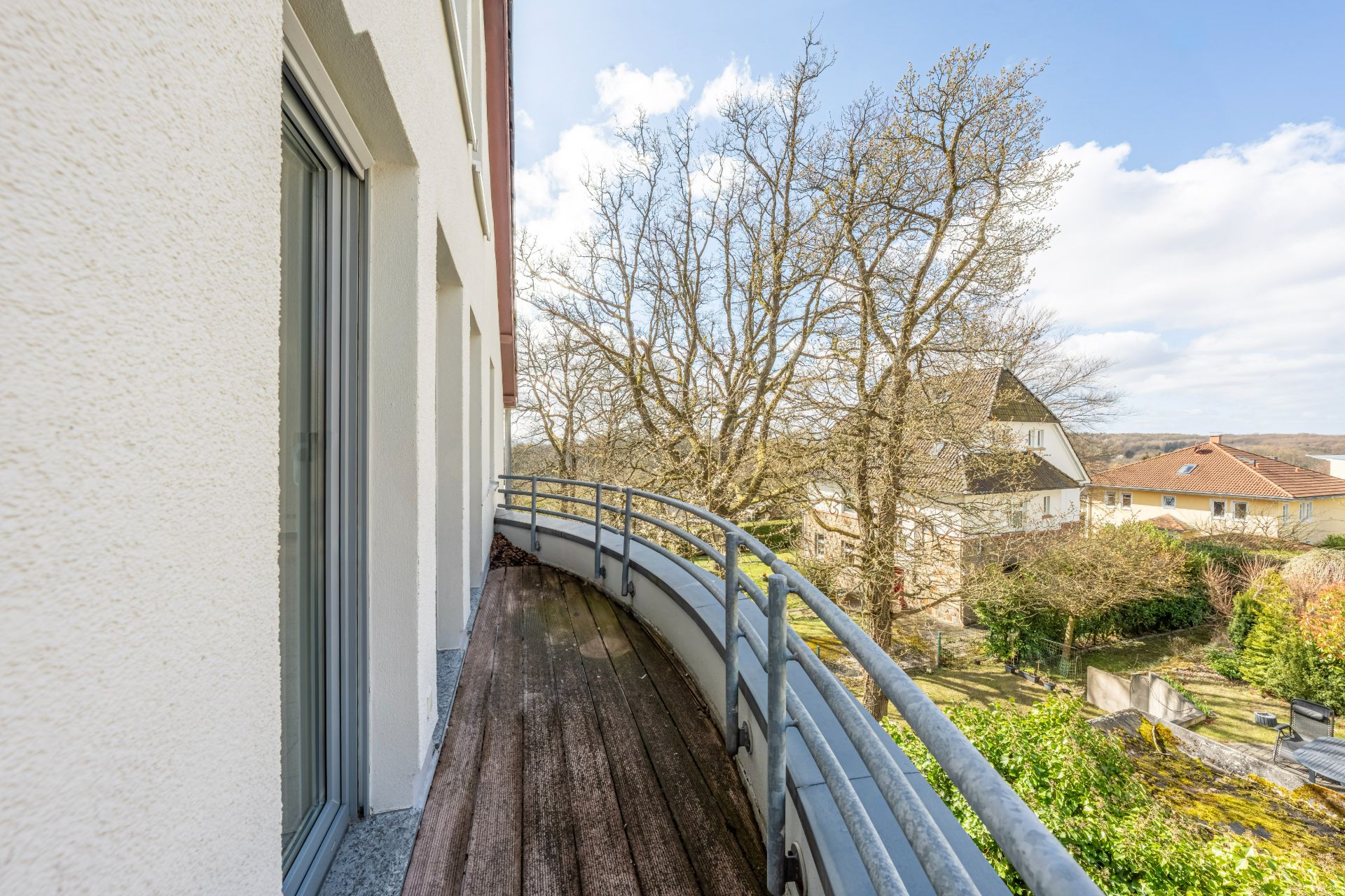 Langer Balkon mit Holzfußboden und Metallgeländer, Blick auf Bäume und Nachbarhäuser.
