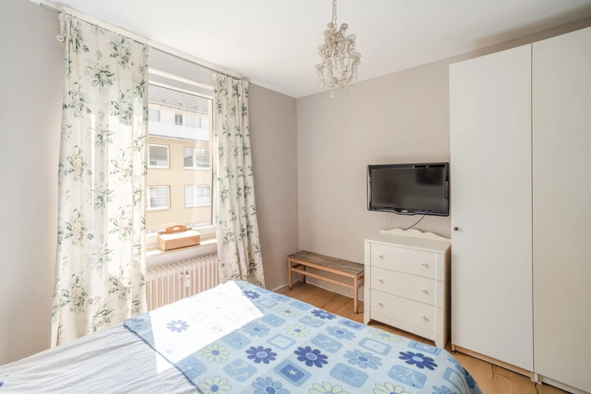 Helles Schlafzimmer mit Bett, Blumentapete, Fernseher, Kommode und Kleiderschrank.
