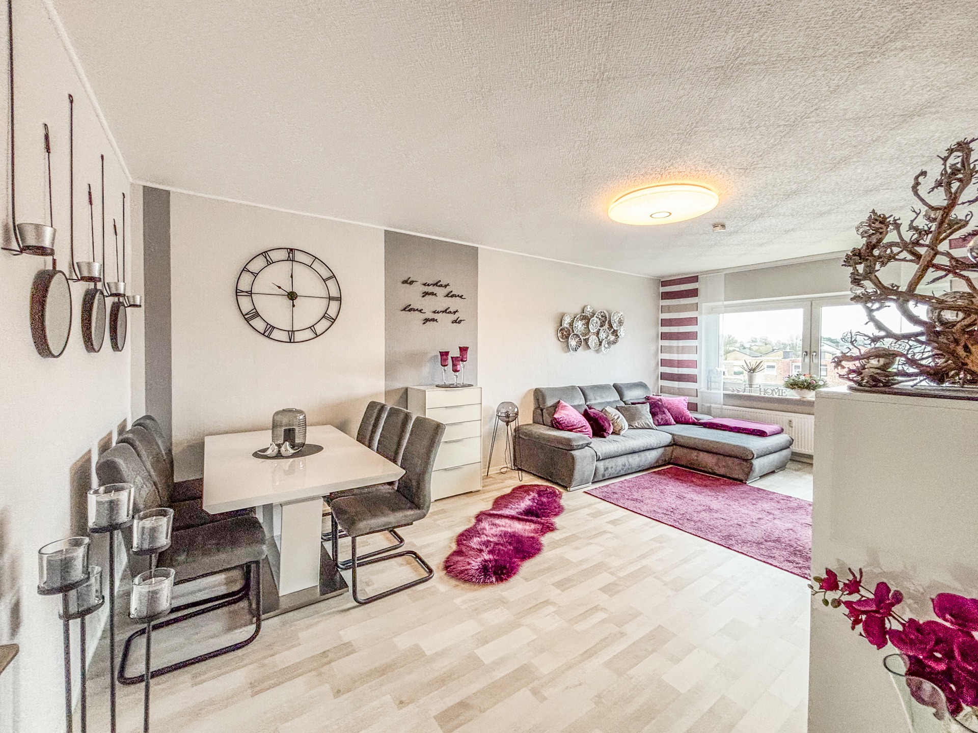 Helles Wohnzimmer mit Essecke, grauem Sofa, pinken Teppichen und großer Wanduhr.