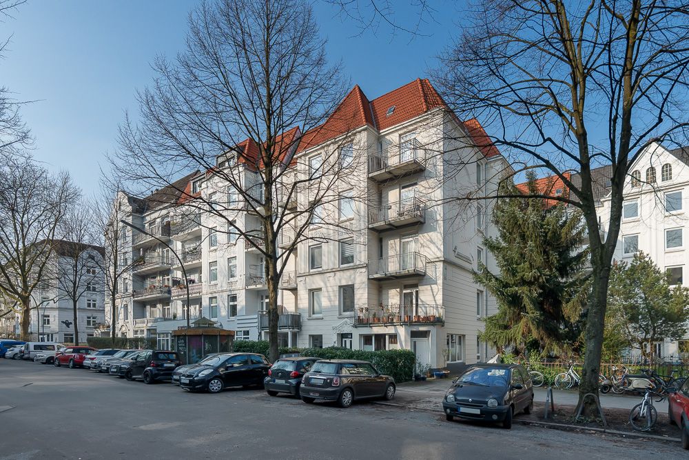 Mehrstöckiges Wohnhaus mit Balkonen, geparkten Autos und Bäumen an der Straße.