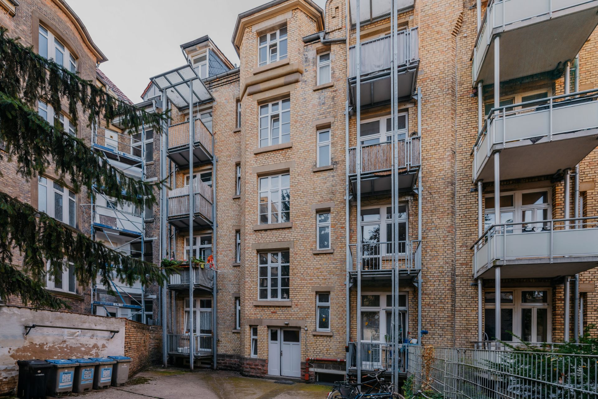 Mehrstöckiges Wohnhaus aus Backstein mit mehreren Balkonen und Fenstern, Hof mit Mülltonnen und Fahrrädern.