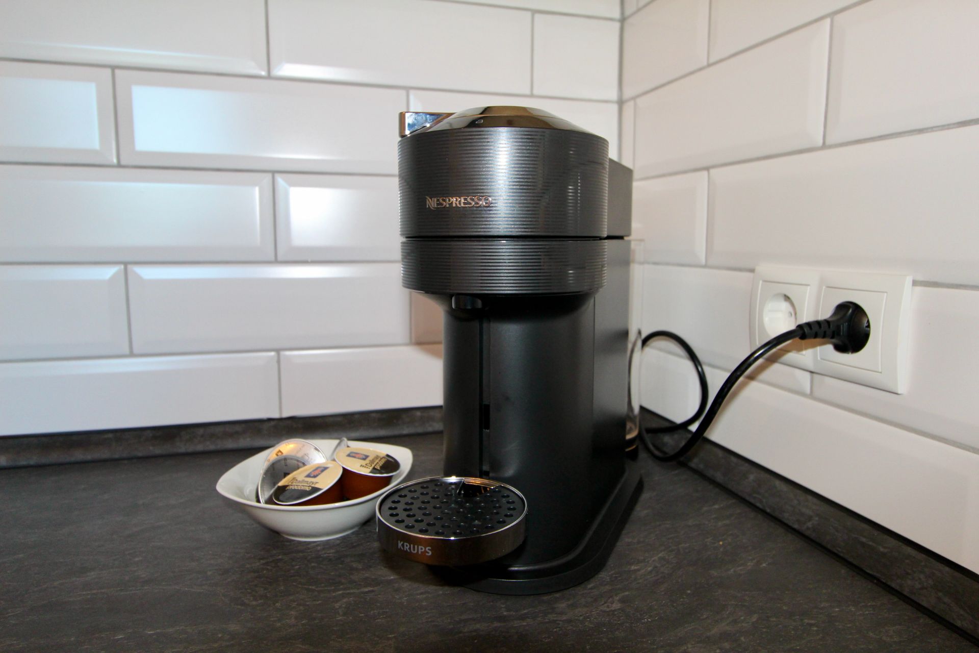 Kaffeemaschine und Schale mit Kaffeekapseln auf Küchentheke vor weißem Fliesenspiegel.