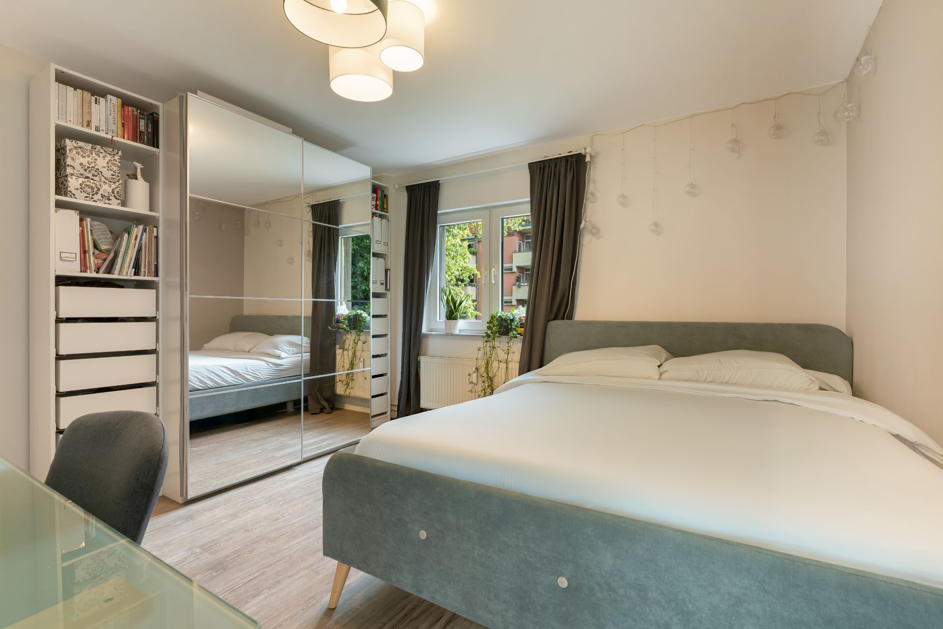Helles Schlafzimmer mit großem Bett, Kleiderschrank mit Spiegeltüren, Regal und Fenster mit Vorhängen.