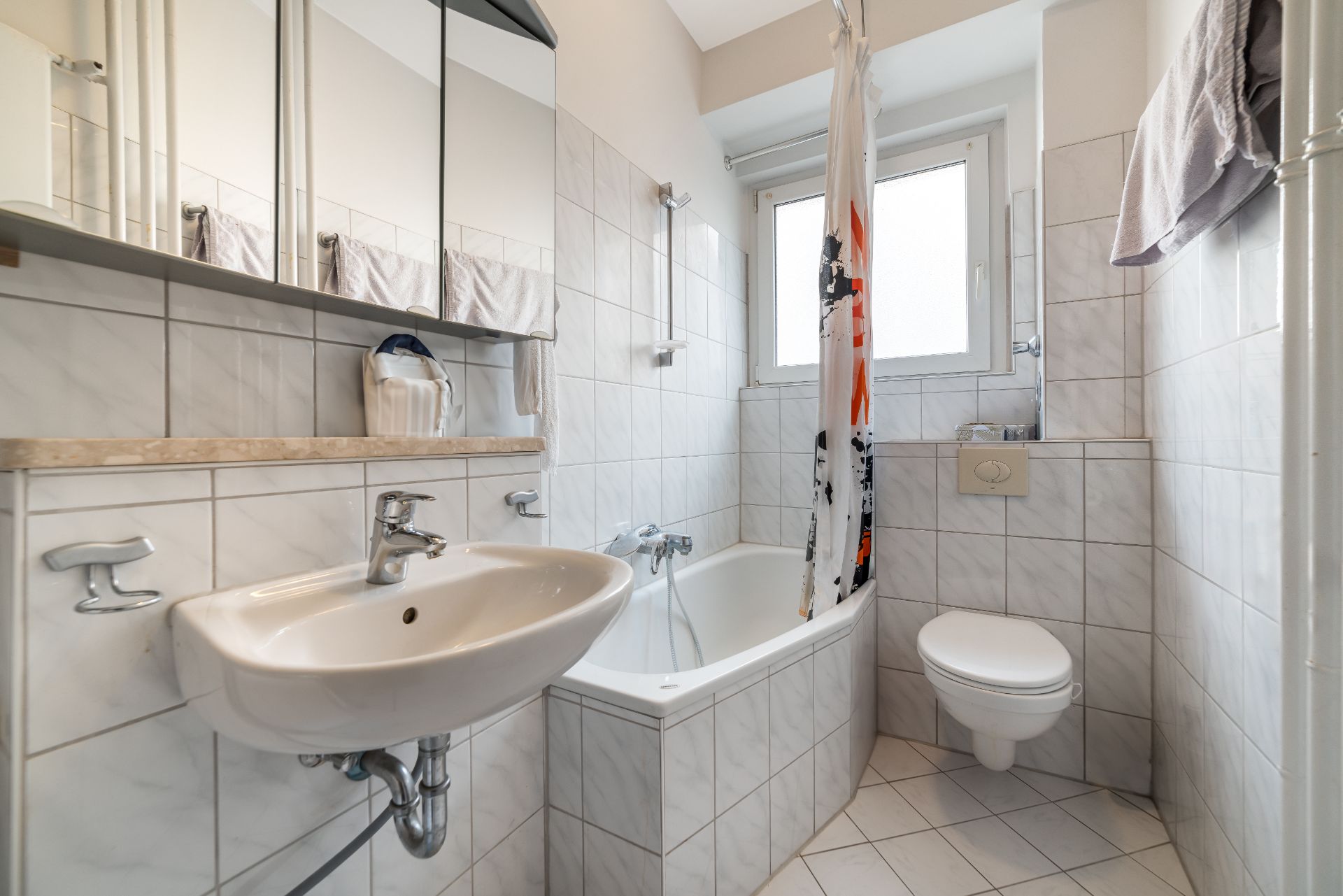 Helles Badezimmer mit Waschbecken, Badewanne, Duschvorhang, WC und Spiegelschrank.