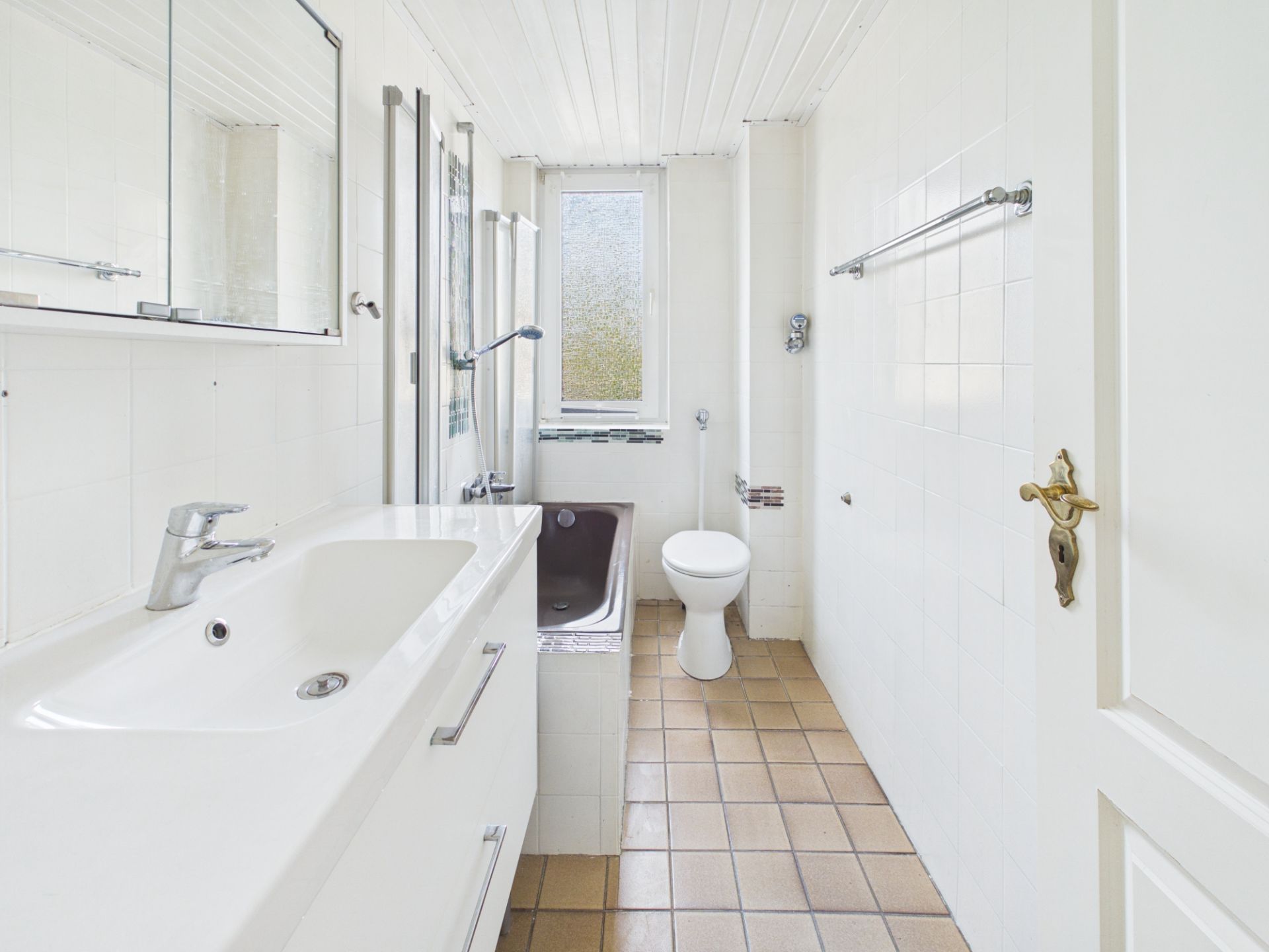 Helles Bad mit Waschbecken, Spiegel, Badewanne und Toilette, brauner Fliesenboden.
