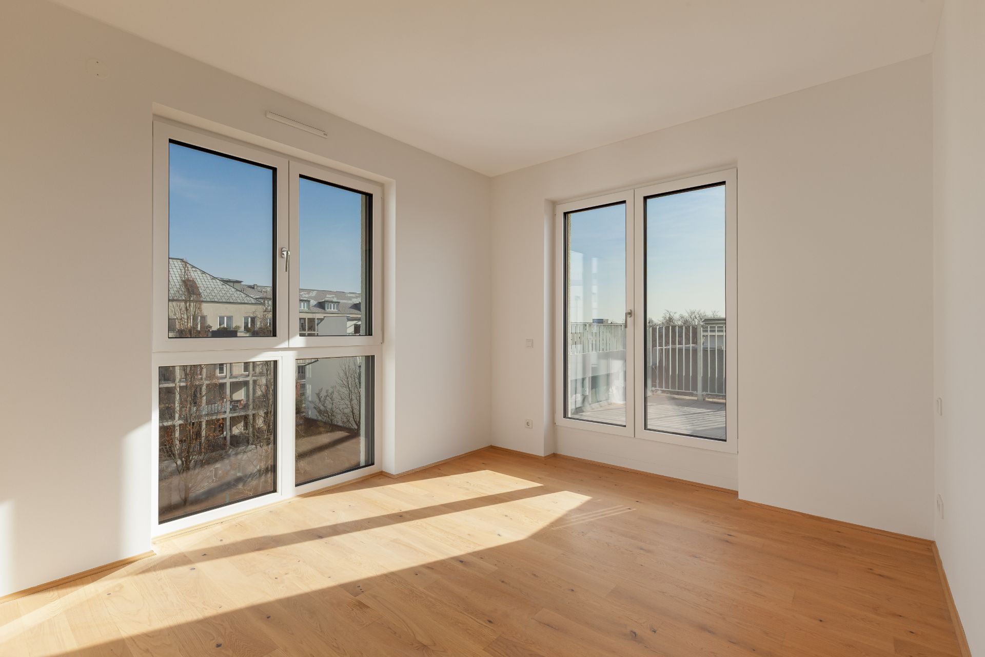 Heller Raum mit großen Fenstern, Holzboden und Blick auf einen Balkon.