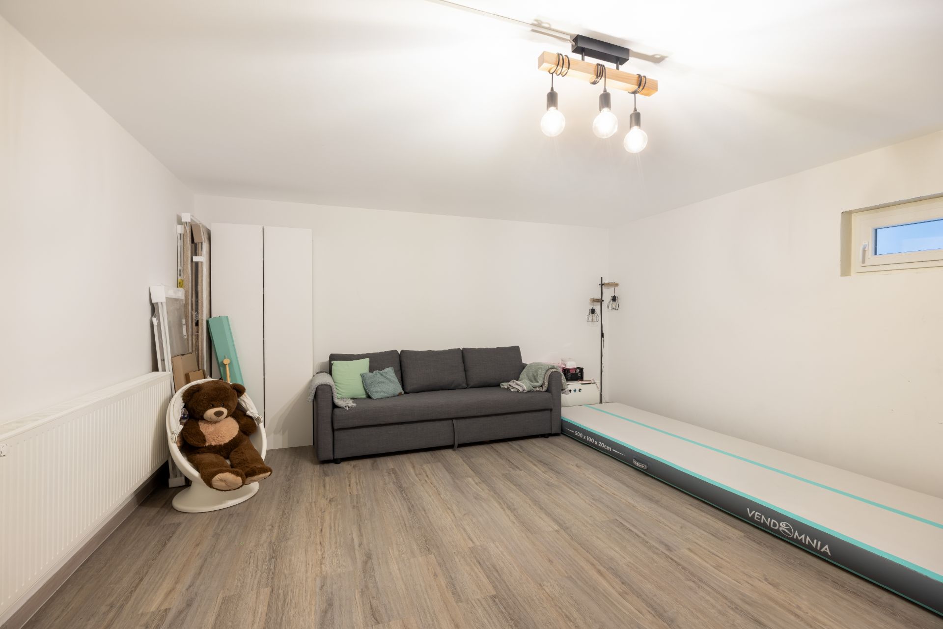 Helles Zimmer mit grauem Sofa, Sessel mit großem Teddybär und Trainingsmatte auf Holzfußboden.