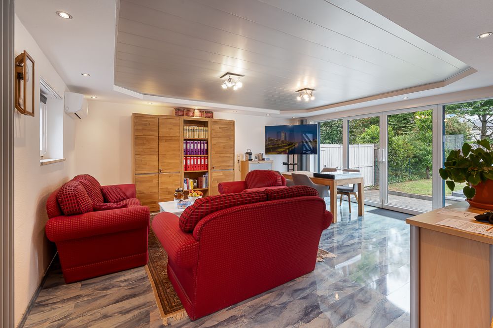 Wohnzimmer mit roten Sofas, Bücherregal, großem Fernseher und Fensterfront mit Blick auf den Garten.