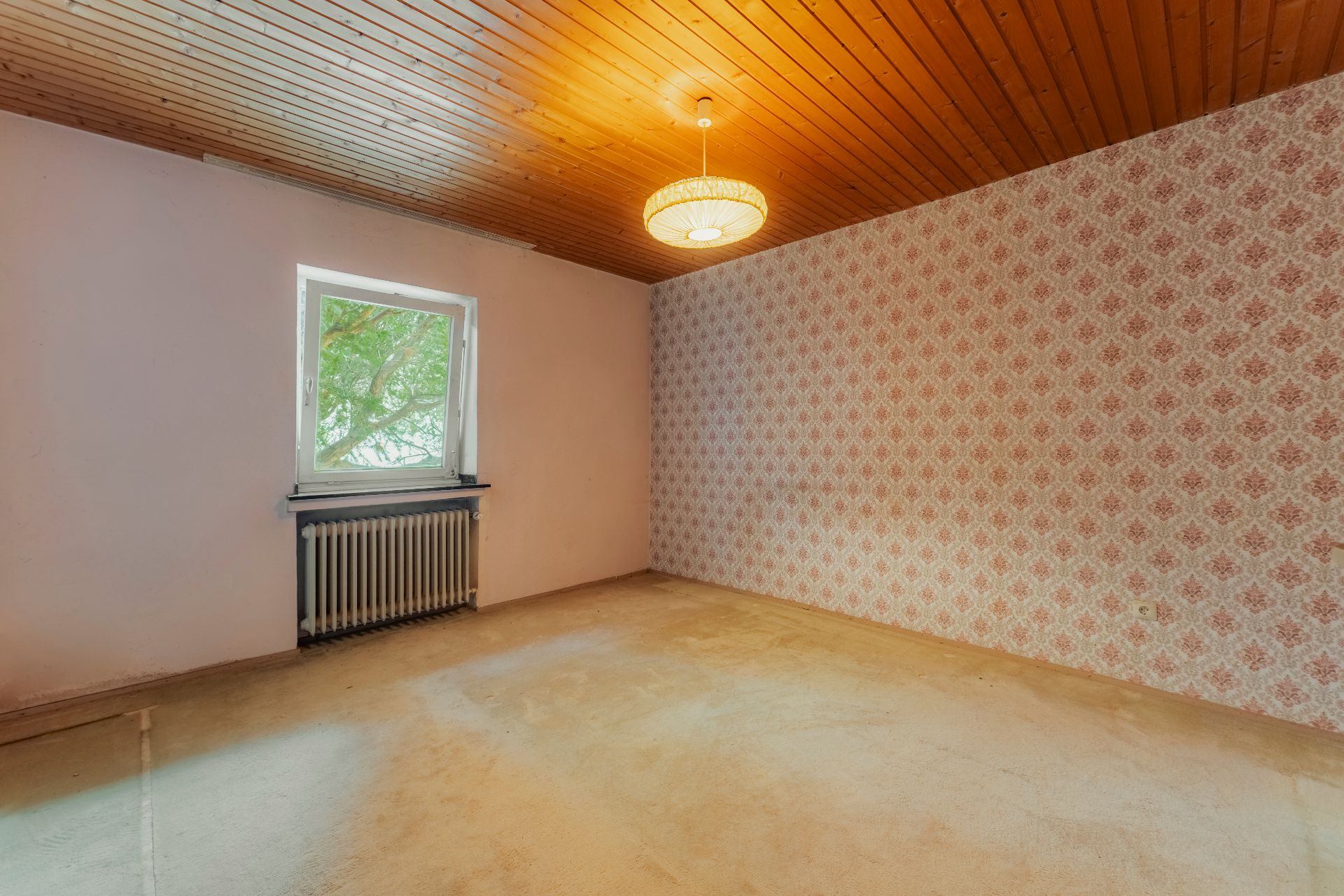 Leeres Zimmer mit Holzbalkendecke, Teppichboden, gemusterter Tapete und Fenster mit Heizung.