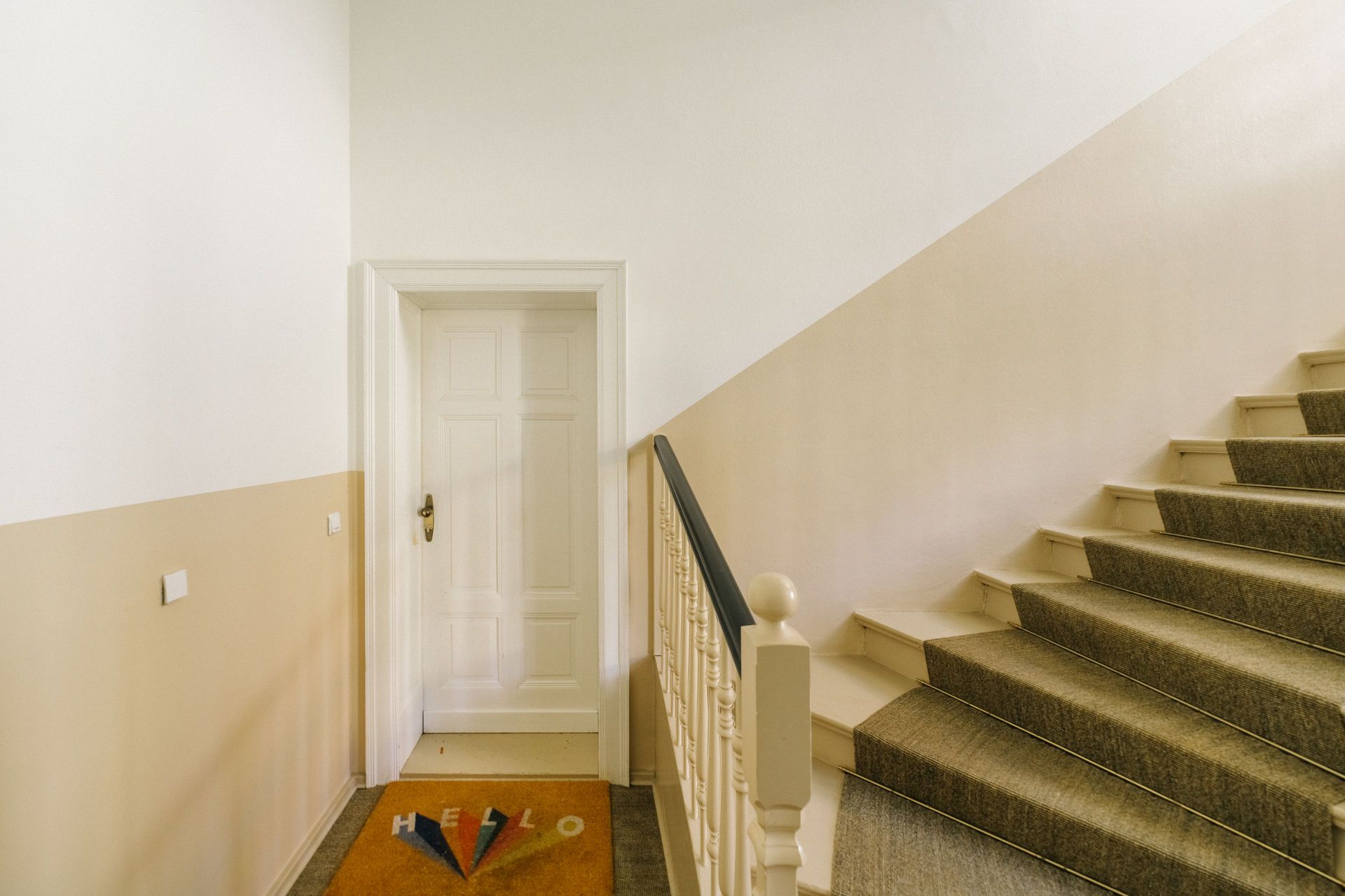 Treppenhaus mit beige gestrichenen Wänden, weißer Tür und Fußmatte mit ‚HELLO‘-Aufschrift.