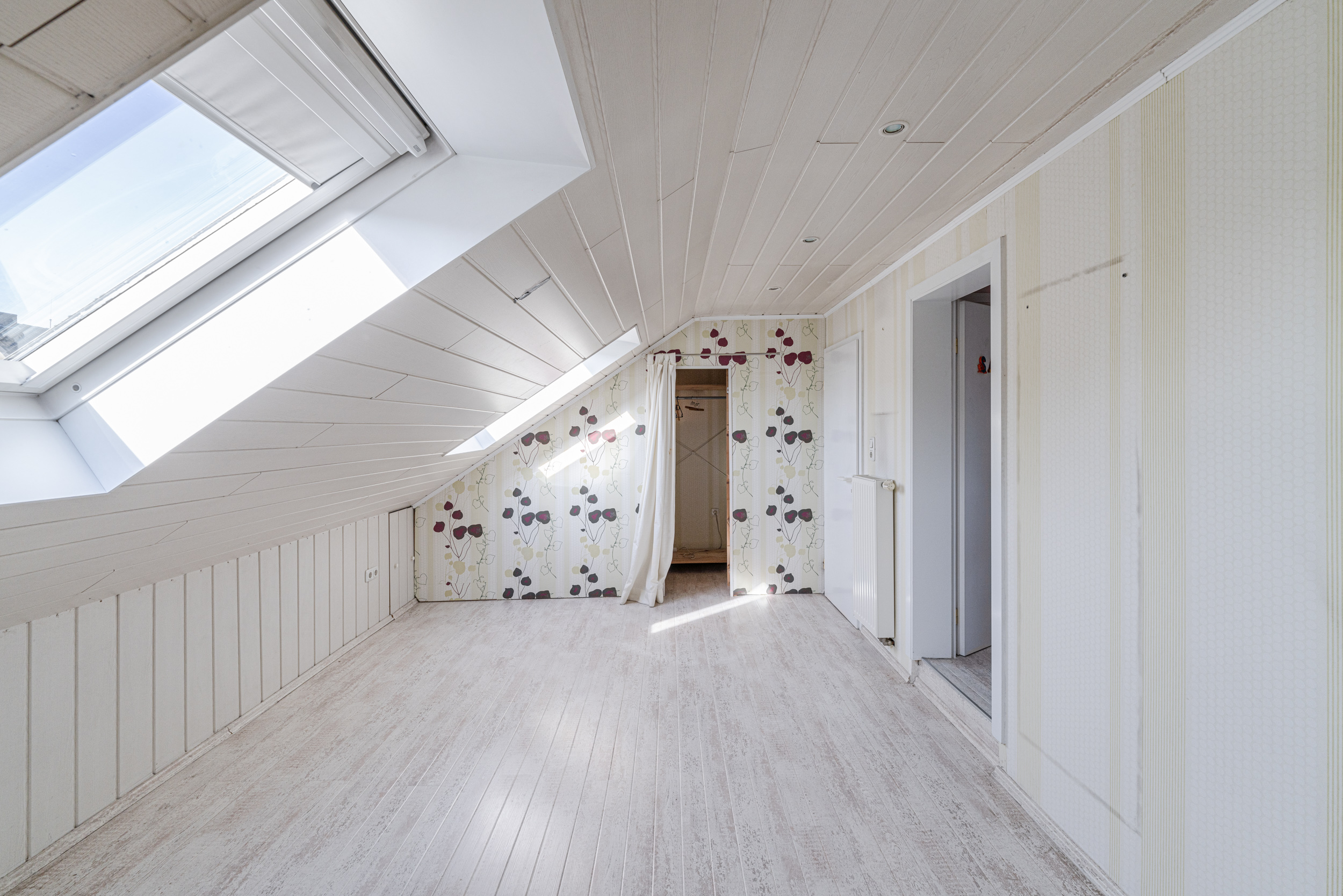 Heller Dachbodenraum mit Dachfenster, Tapete mit Blumenmuster und weißem Holzboden.