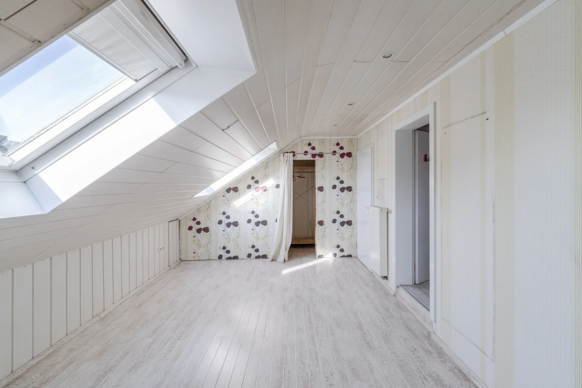 Heller Dachbodenraum mit Dachfenster, Tapete mit Blumenmuster und weißem Holzboden.