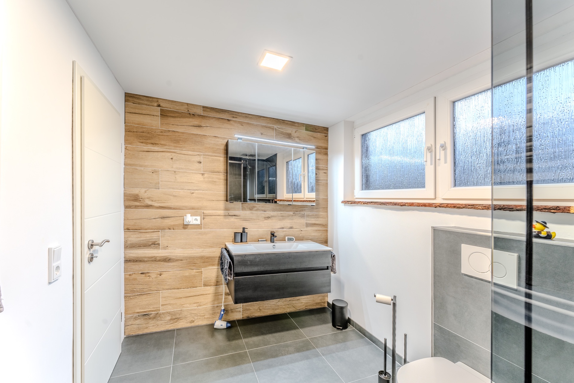 Modernes Badezimmer mit Holzverkleidung, großem Spiegel, Waschbecken, Toilette und großen Fenstern.