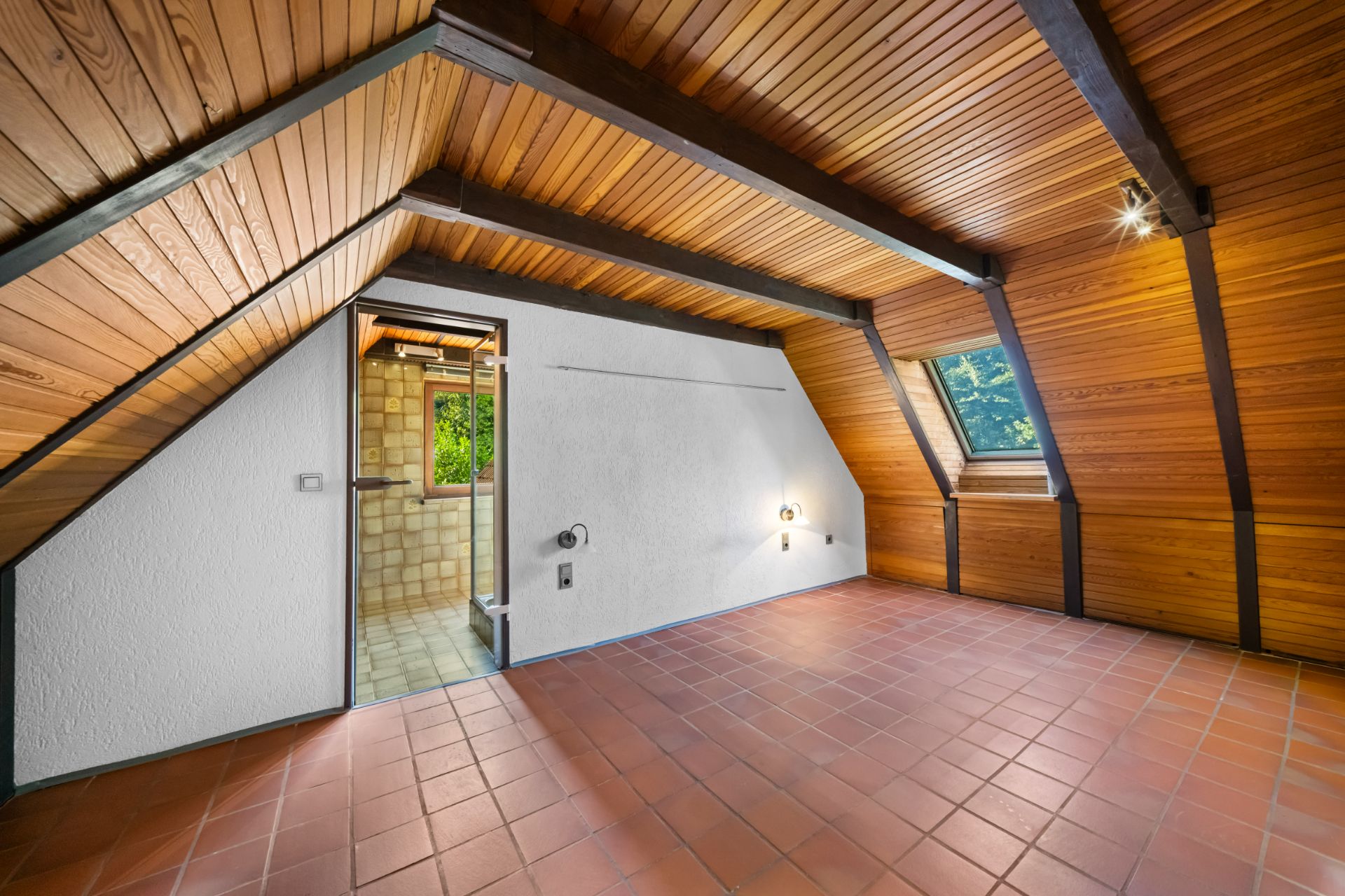 Dachzimmer mit Holzdecke, Fliesenboden, schrägen Wänden und Fenster, Tür führt in gefliestes Bad.