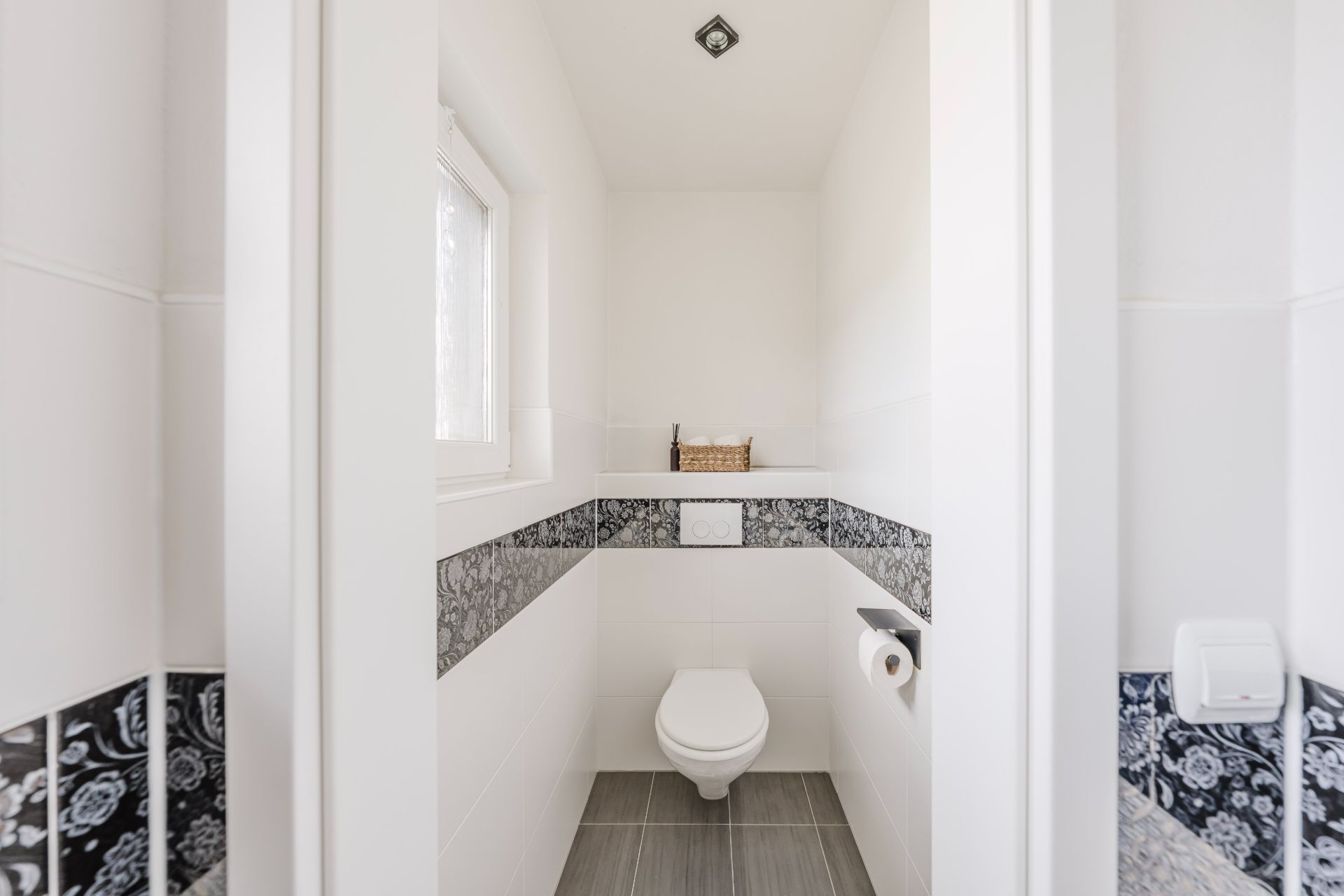 Helles, schmales Badezimmer mit Wandtoilette, Fenster und gemusterten Fliesen.