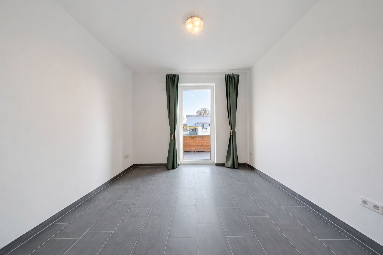 Leerstehender Raum mit grauem Fliesenboden, Türen und grünen Vorhängen vor Fenster.