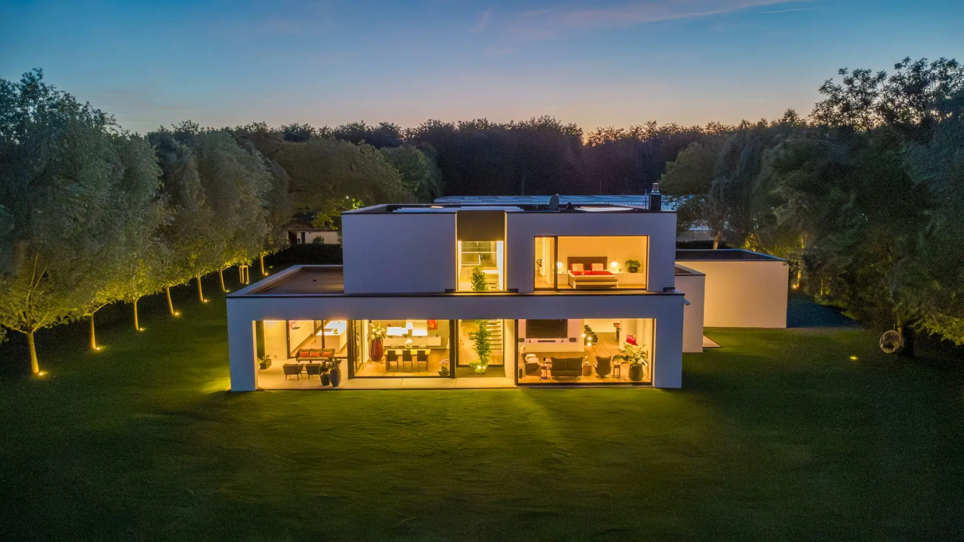 Modernes, zweistöckiges Wohnhaus mit großen Fenstern, beleuchtet im Garten bei Dämmerung.