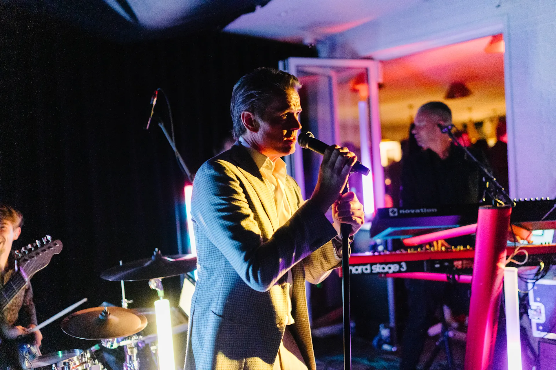 Mehrere Musiker spielen in einem Wohnzimmer, ein Mann singt am Mikrofon, bunte Lichter leuchten.