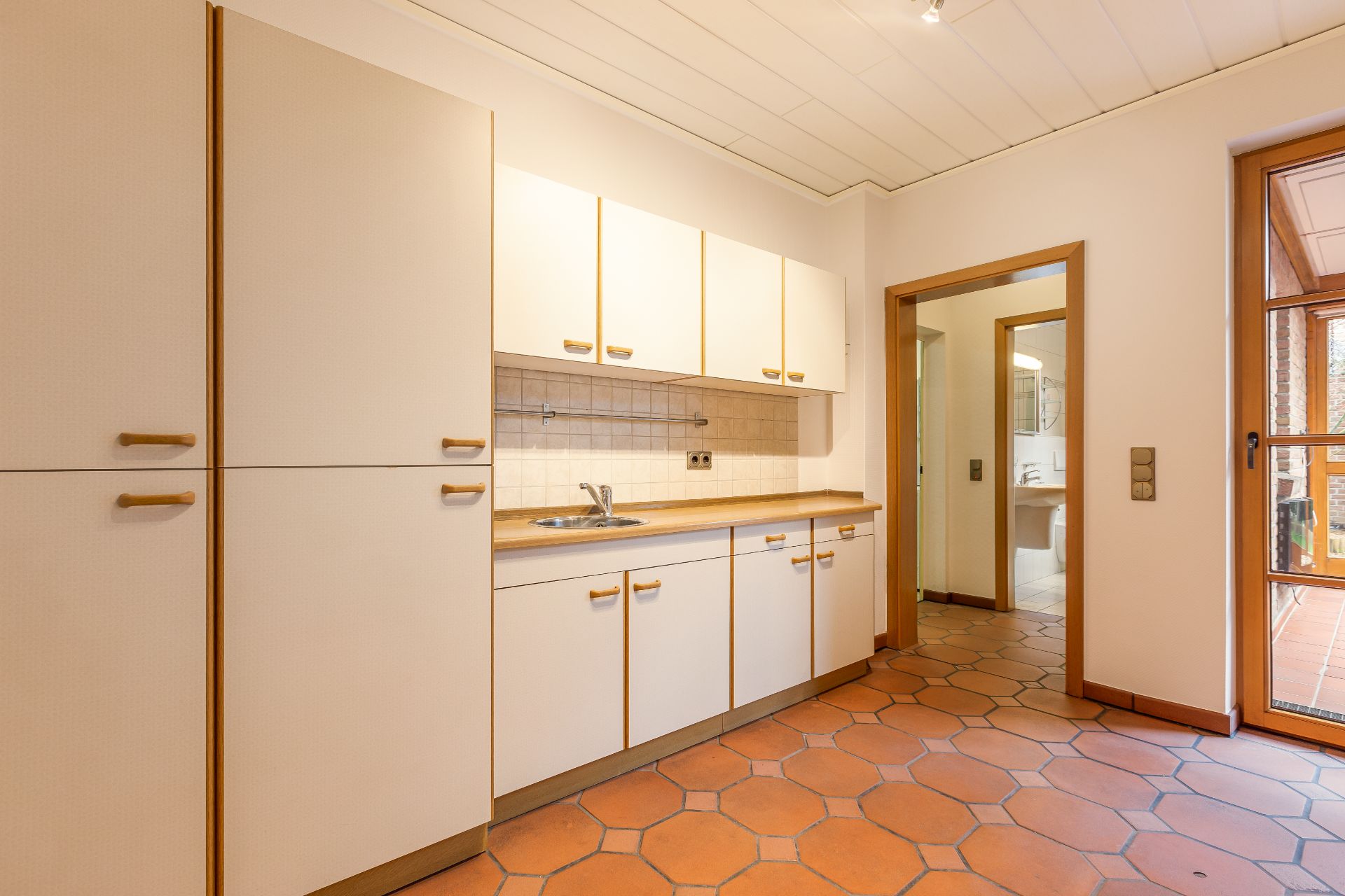 Helle Küche mit Einbauschränken, Spüle und Fliesenboden, Tür zum Flur und Fenster zur Terrasse.