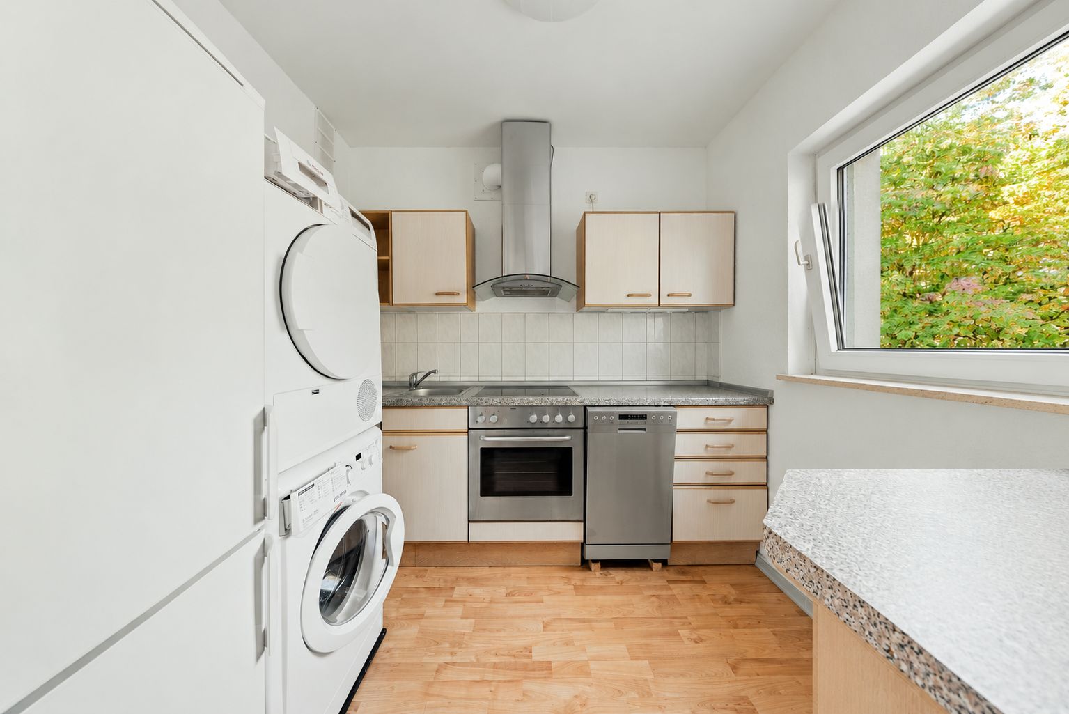 Moderne Küche mit Waschmaschine, Herd, Spülmaschine und großem Fenster mit Blick ins Grüne.