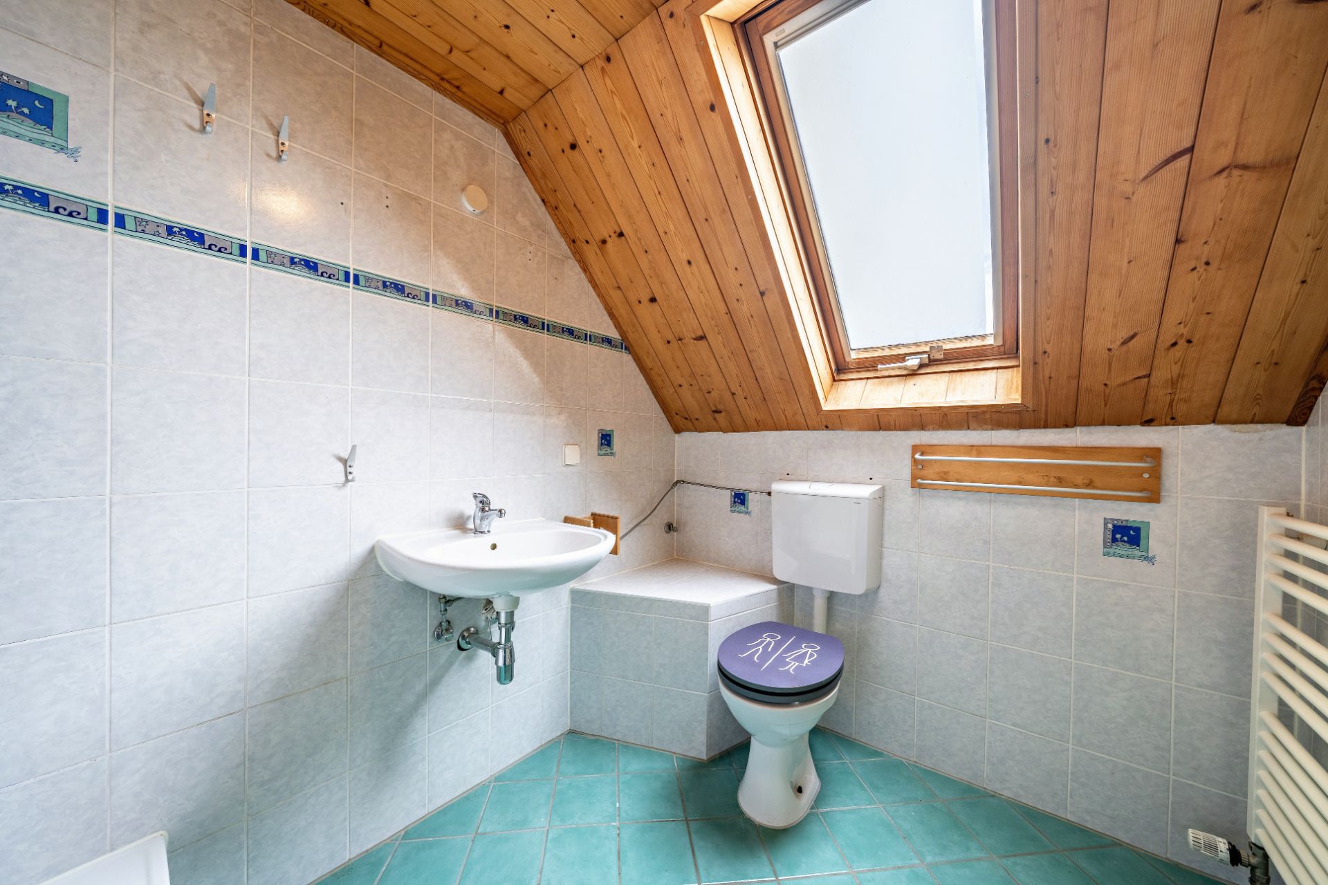 Helles Bad mit Dachfenster, Waschbecken, Toilette, Holzdecke und türkisfarbenen Fliesen.