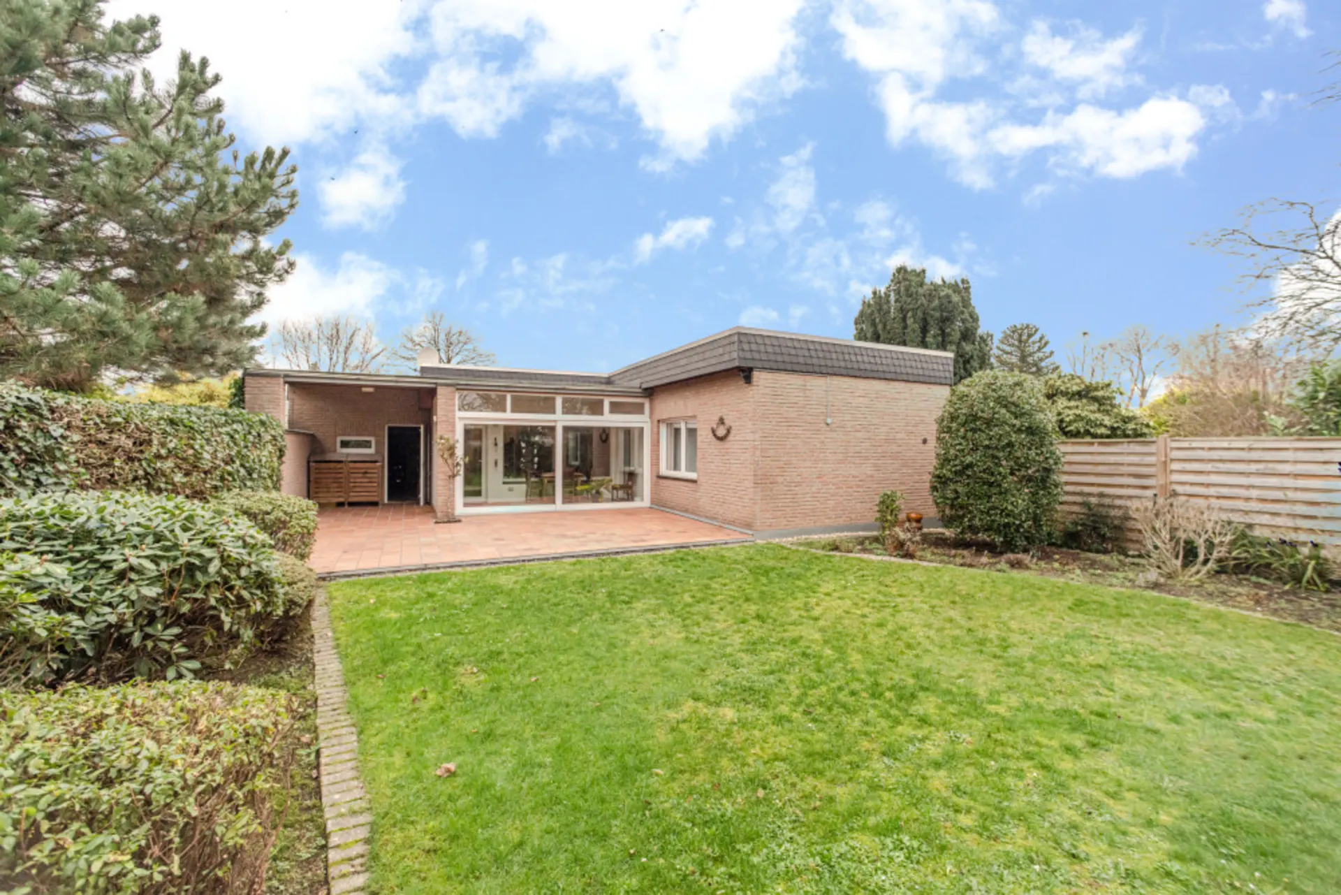 Backsteinbungalow mit großer Terrasse und gepflegtem Garten, umgeben von Hecken und Zaun.