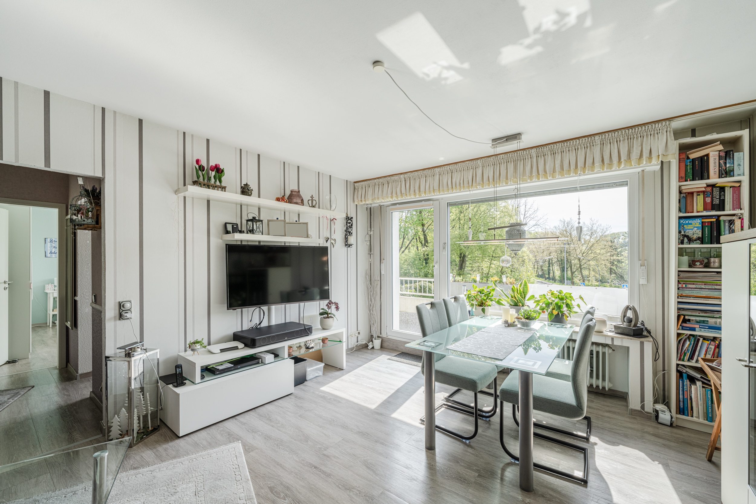 Helles Wohnzimmer mit Esstisch, Glasplatte, Stühlen, TV, Bücherregal und großen Fenstern.