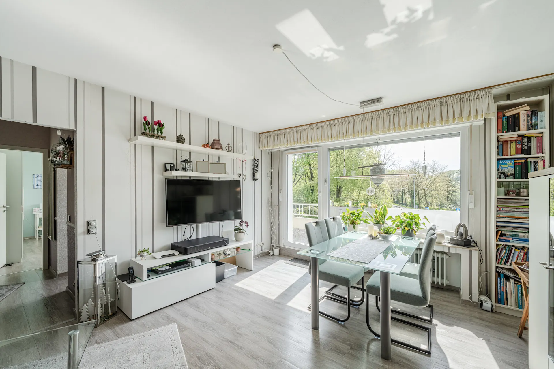 Helles Wohnzimmer mit Esstisch, Glasplatte, Stühlen, TV, Bücherregal und großen Fenstern.