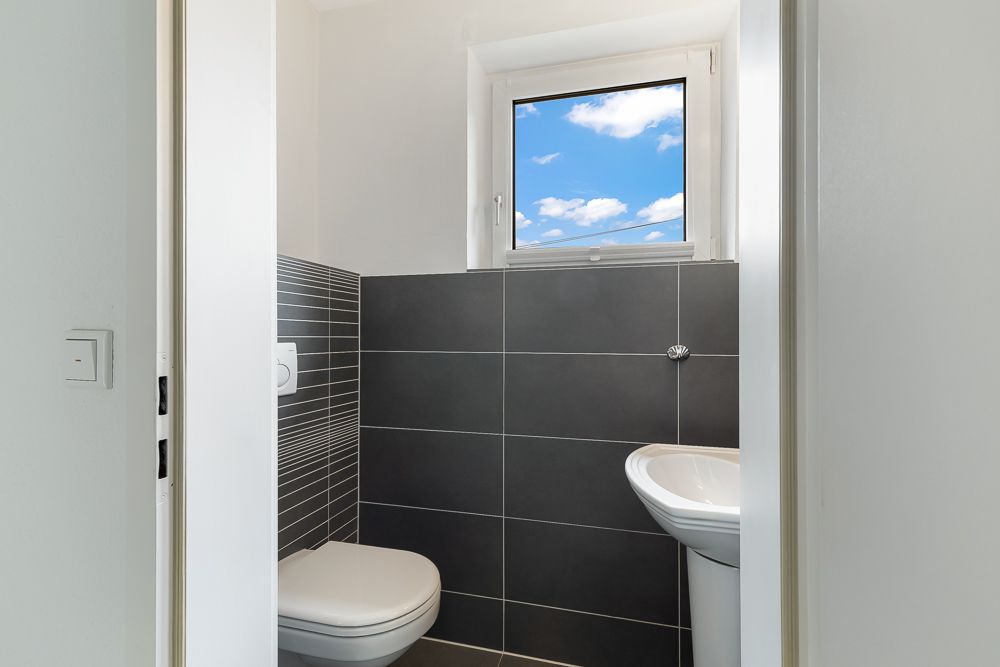 Kleines Bad mit Toilette, Waschbecken, grauen Fliesen und Fenster.