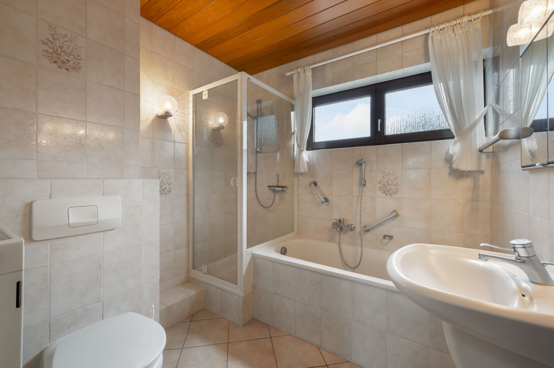 Helles Badezimmer mit Badewanne, Dusche, Waschbecken, Toilette und Fenster mit Vorhängen.