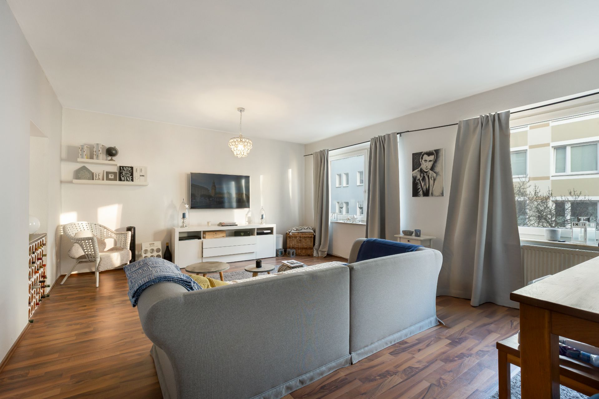 Helles Wohnzimmer mit großer grauer Couch, Holzboden, TV an der Wand und zwei großen Fenstern mit Vorhängen.