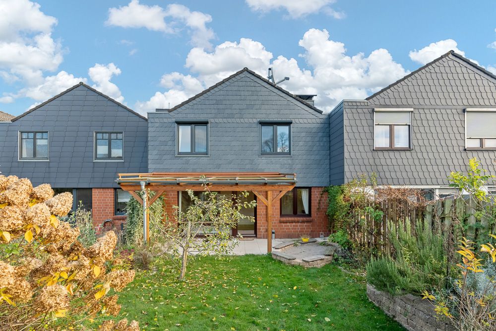 Reihenhaus mit Garten, Terrasse und Überdachung, umgeben von Pflanzen und Büschen.