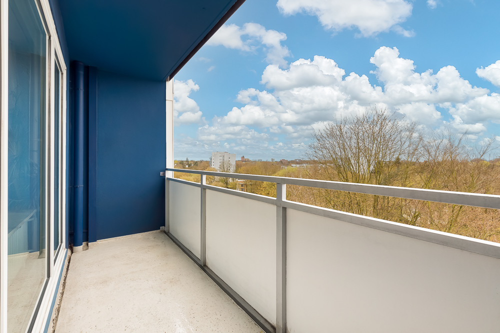 Balkon mit blau gestrichener Wand, Glasgeländer und Ausblick auf Bäume und Himmel.