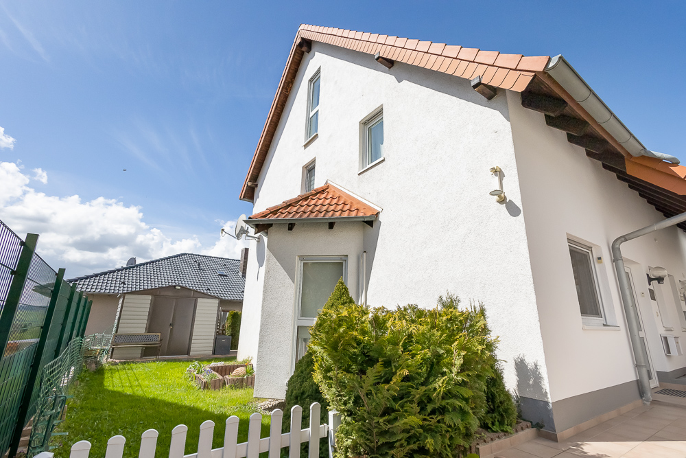 Weißes Einfamilienhaus mit rotem Dach, Garten, Zaun und Sträuchern im Vordergrund.