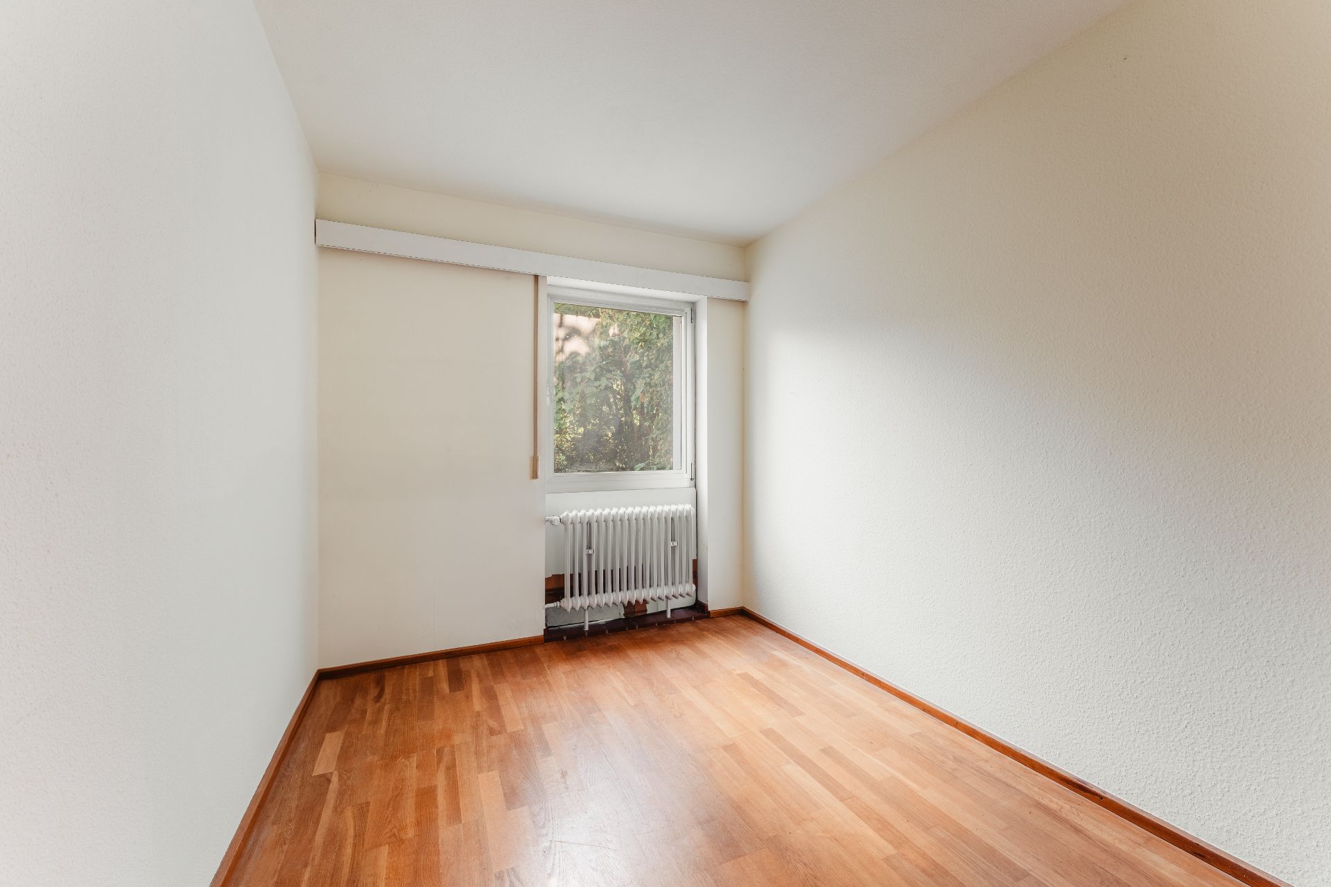 Leerer Raum mit Holzfußboden, weiß gestrichenen Wänden und einem Fenster mit Heizkörper.
