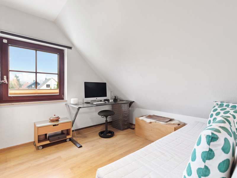 Helles Zimmer mit Holzfußboden, Schreibtisch, Computer, Stuhl, Bett und Fenster.